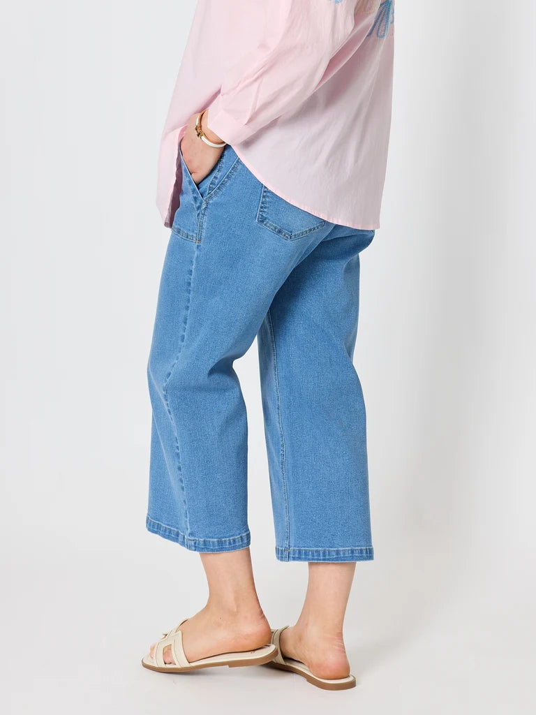 A woman wearing Gordon Smith light blue cropped denim jeans, a light pink long-sleeve top, and beige slide sandals, shown from the waist down against a plain light gray studio background.