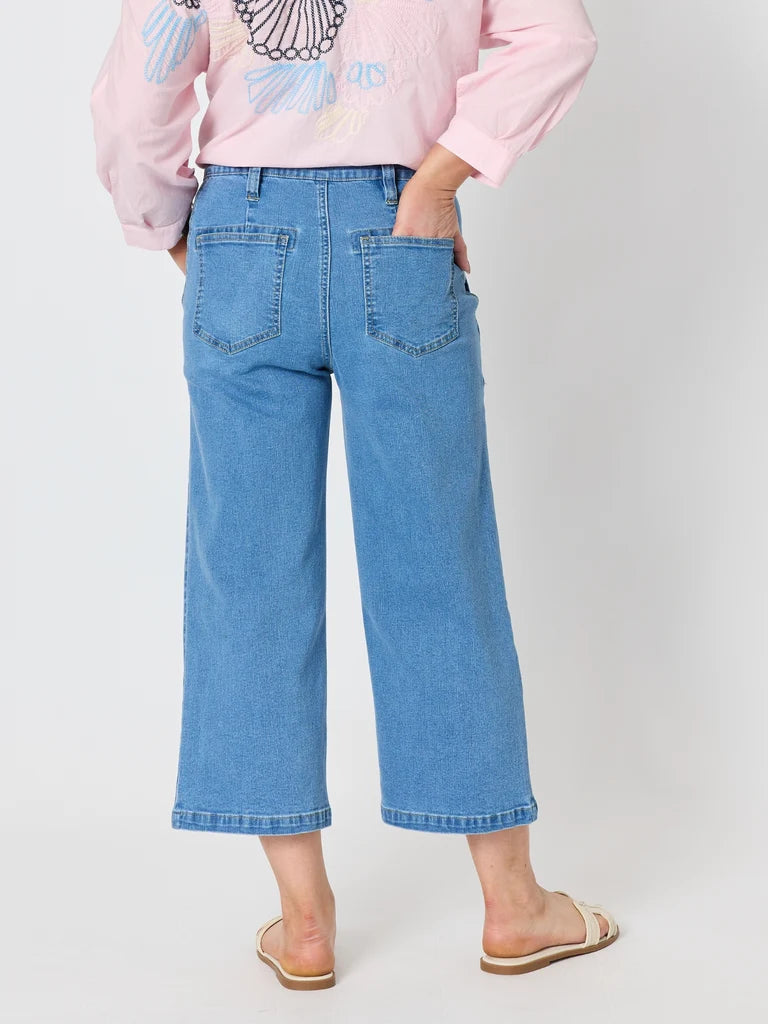 A model shown from the back wearing Gordon Smith light blue cropped wide-leg denim jeans, a pink long-sleeve top, and beige slide sandals, set against a plain light gray studio background.