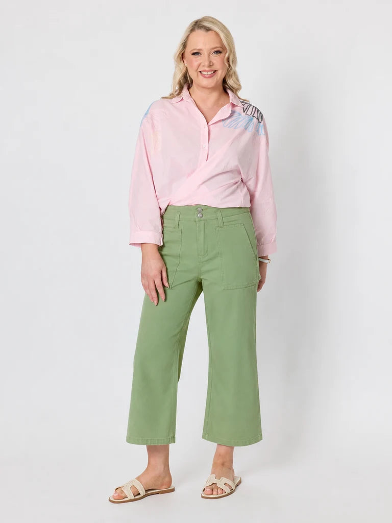 Image of A woman modeling a light pink long-sleeve button-up shirt with abstract blue and purple patterns on the shoulders, paired with Gordon Smith sage green cropped pants and beige slide sandals, against a light gray studio background.