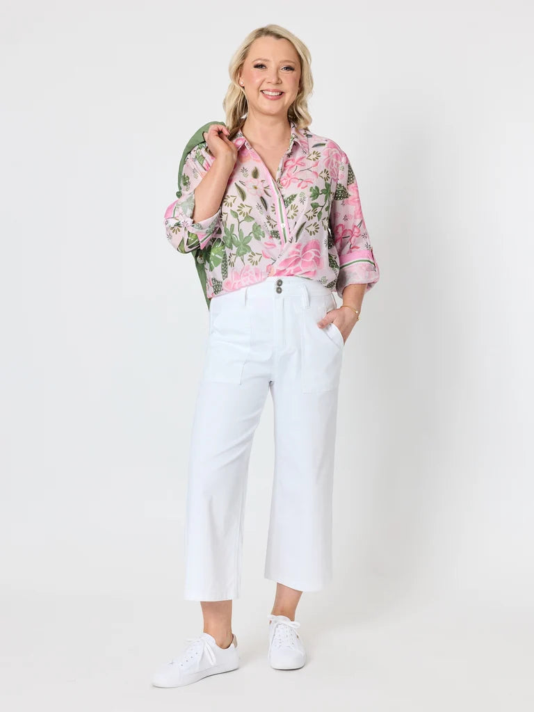 A woman modeling a pink and green floral print button-up shirt paired with Gordon Smith white cropped trousers and white sneakers, standing against a plain white studio background.