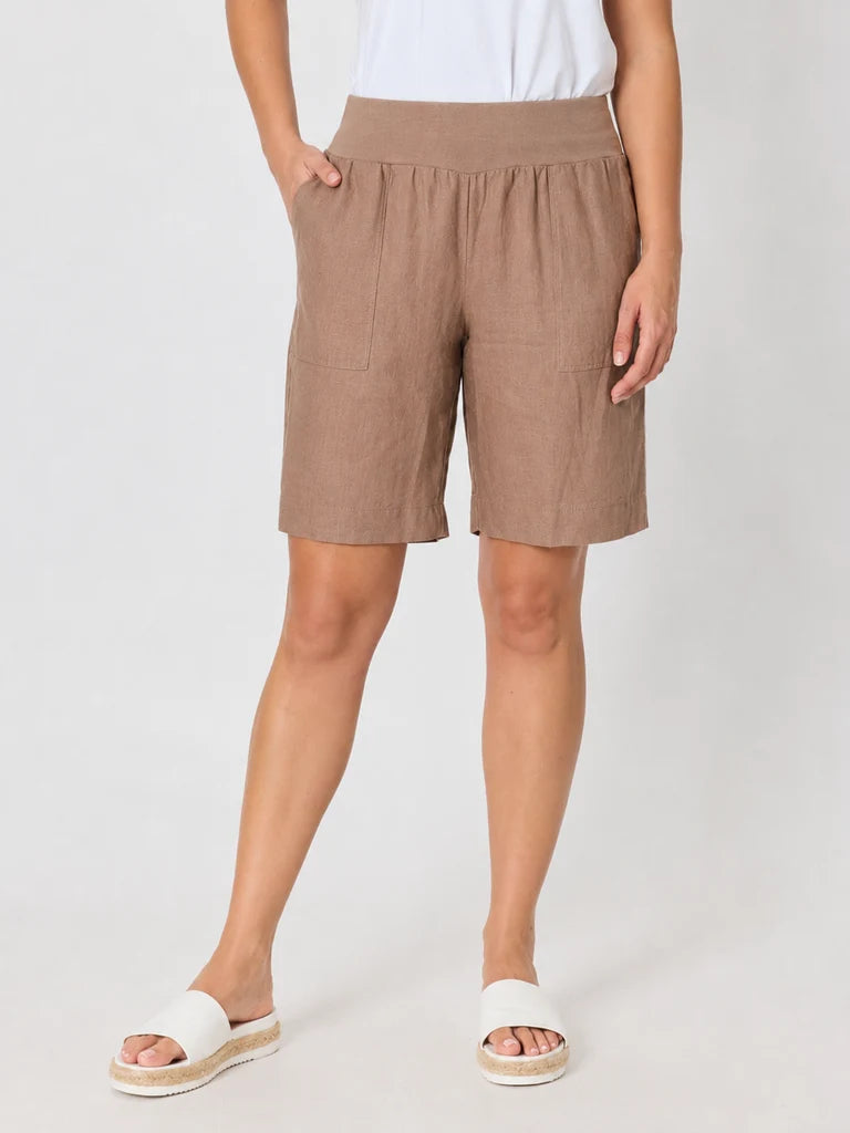 Image of Woman wearing Gordon Smith knee-length taupe linen shorts with side pockets, paired with a white top and white slide sandals, standing against a light gray studio background.