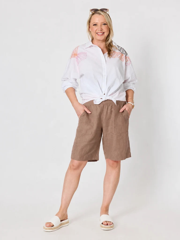 Image of A woman modeling a white long-sleeve button-up shirt with pink and gray butterfly details on the shoulders, paired with Gordon Smith brown linen shorts and white slide sandals, against a light gray studio background.