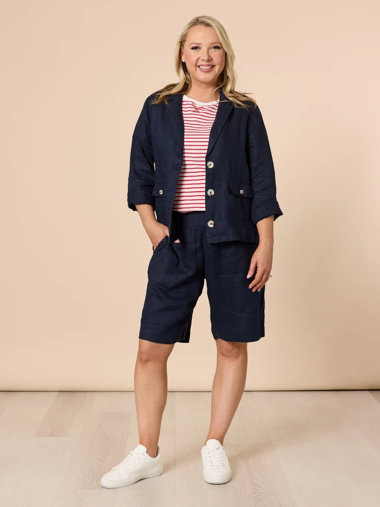 A woman modeling a navy blue linen-blend short jacket and Gordon Smith matching shorts set, styled over a red and white striped t-shirt with white sneakers, against a solid beige studio background.