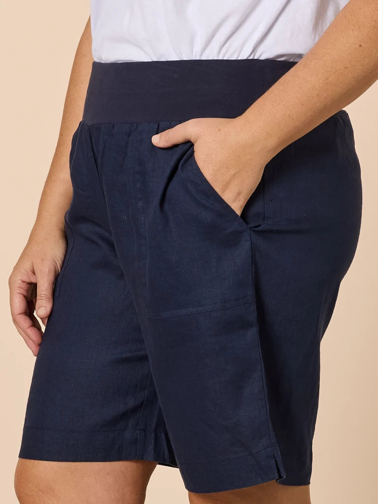 Image of A close-up of a woman wearing Gordon Smith dark navy blue linen shorts with a matching wide waistband, paired with a white top, set against a solid beige background.