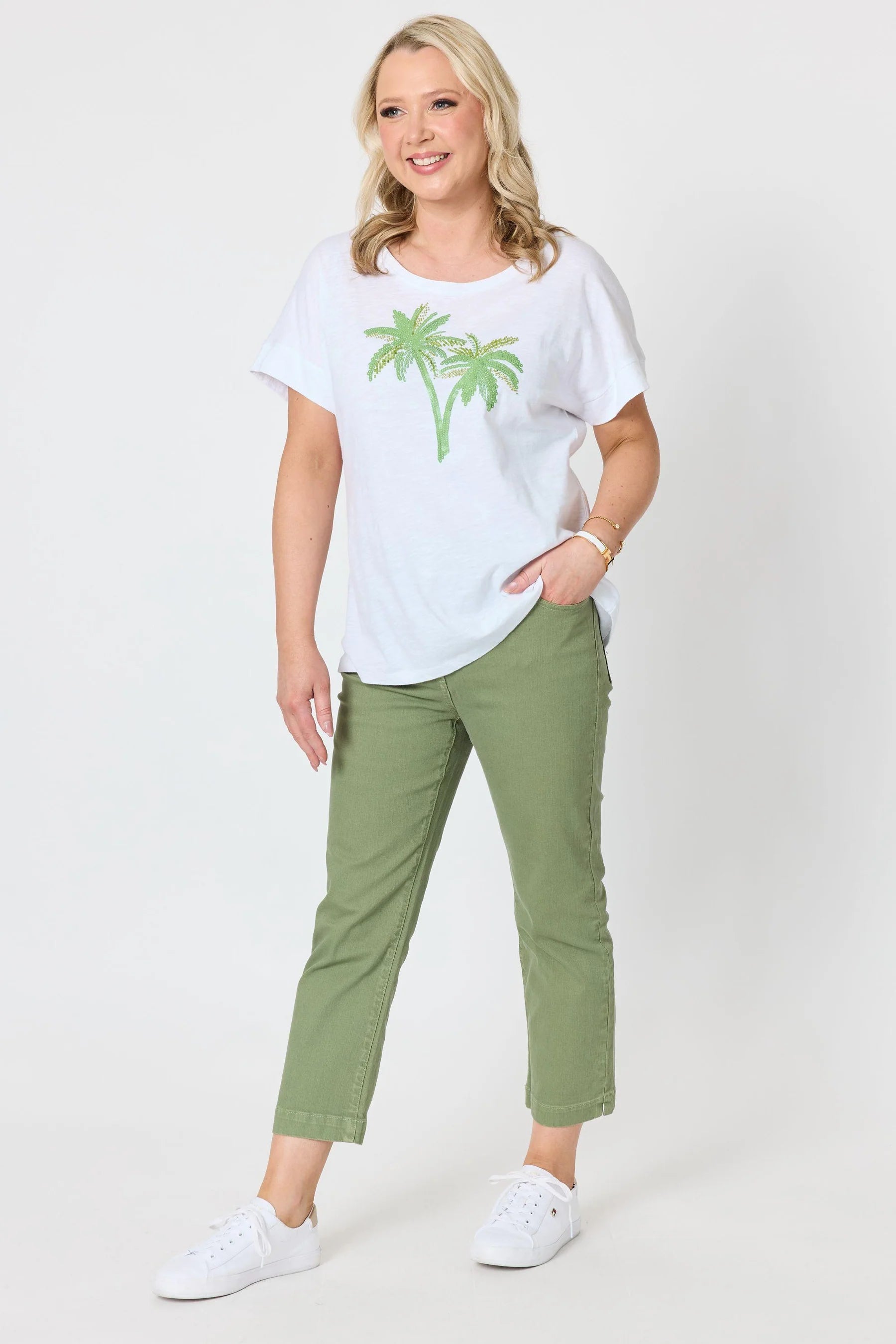 Image of A woman modeling a Gordon Smith white short-sleeve t-shirt with a green palm tree graphic, paired with olive green cropped pants and white sneakers, against a light gray studio background.