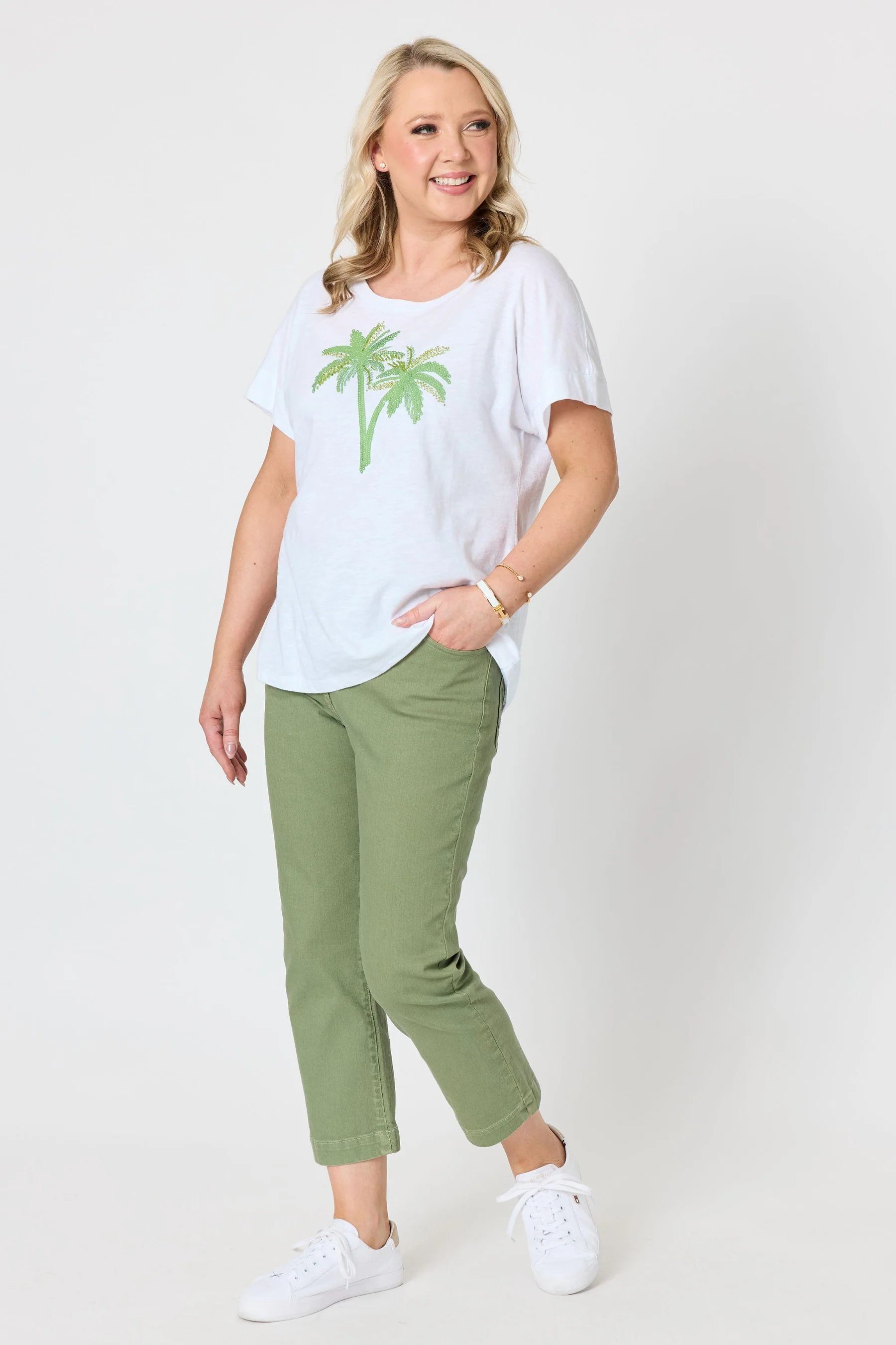 A woman modeling a Gordon Smith white short-sleeve t-shirt with a green palm tree graphic, paired with olive green cropped pants and white sneakers, against a light gray studio background.