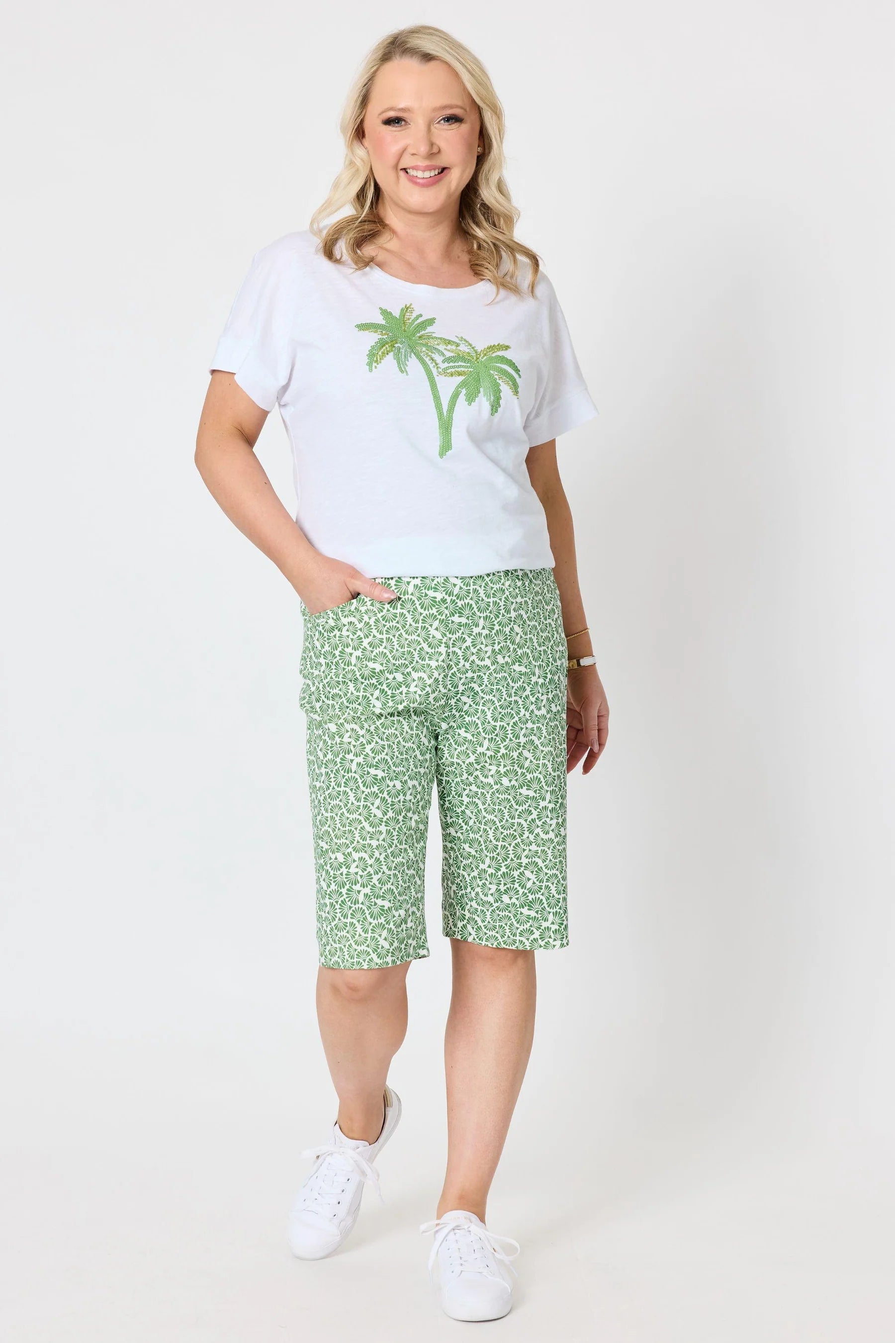 A woman modeling a Gordon Smith white short-sleeve t-shirt with a green palm tree graphic and green patterned knee-length shorts, paired with white sneakers, against a plain white studio background.
