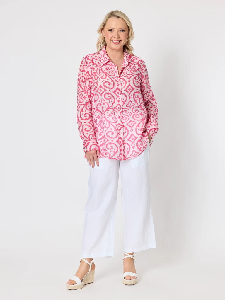 Image of A woman modeling a Gordon Smith pink and white patterned button-down shirt with white cropped pants and white wedge sandals, standing against a plain white studio background.