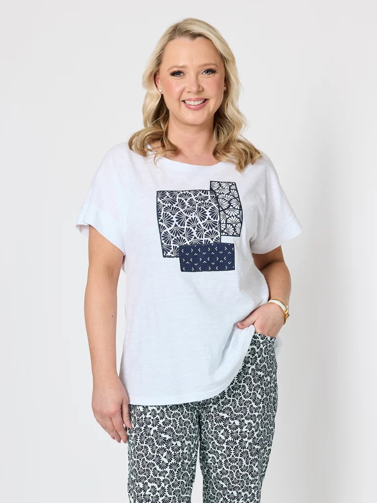 A woman modeling a Gordon Smith white short-sleeve t-shirt with a navy blue geometric patchwork print on the front, paired with matching navy and white patterned pants, against a light gray studio background.