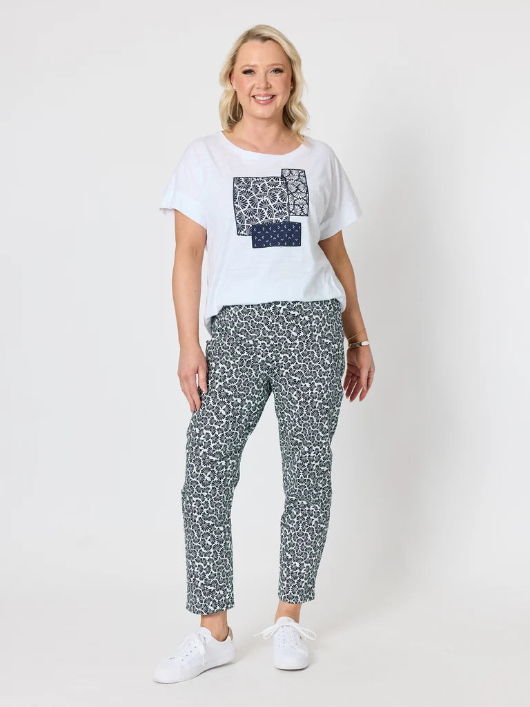 Image of A woman modeling a Gordon Smith white graphic t-shirt and navy blue patterned cropped trousers with white sneakers, standing against a light gray studio background.