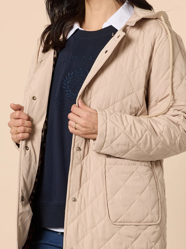 Woman wearing a Gordon Smith beige quilted coat over a navy sweater with a logo on a beige background