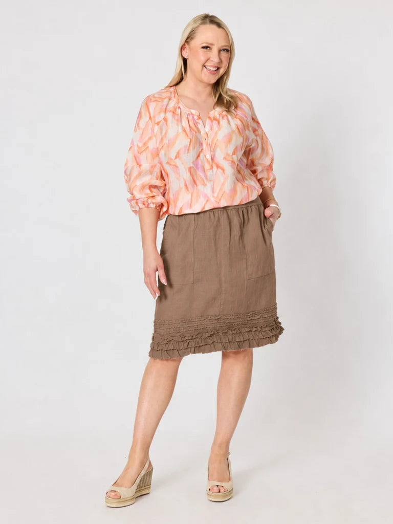 Image of A woman modeling a peach and orange abstract print blouse with three-quarter puffed sleeves, paired with a Gordon Smith brown ruffled knee-length skirt and beige wedge sandals, against a light gray studio background.
