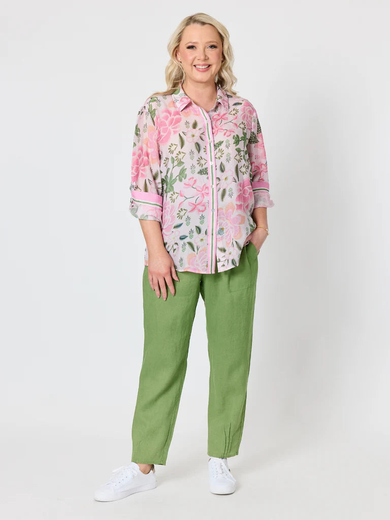 Image of A woman modeling a pink floral button-up shirt with green and white details, paired with Gordon Smith olive green pants and white sneakers, against a light gray studio background.