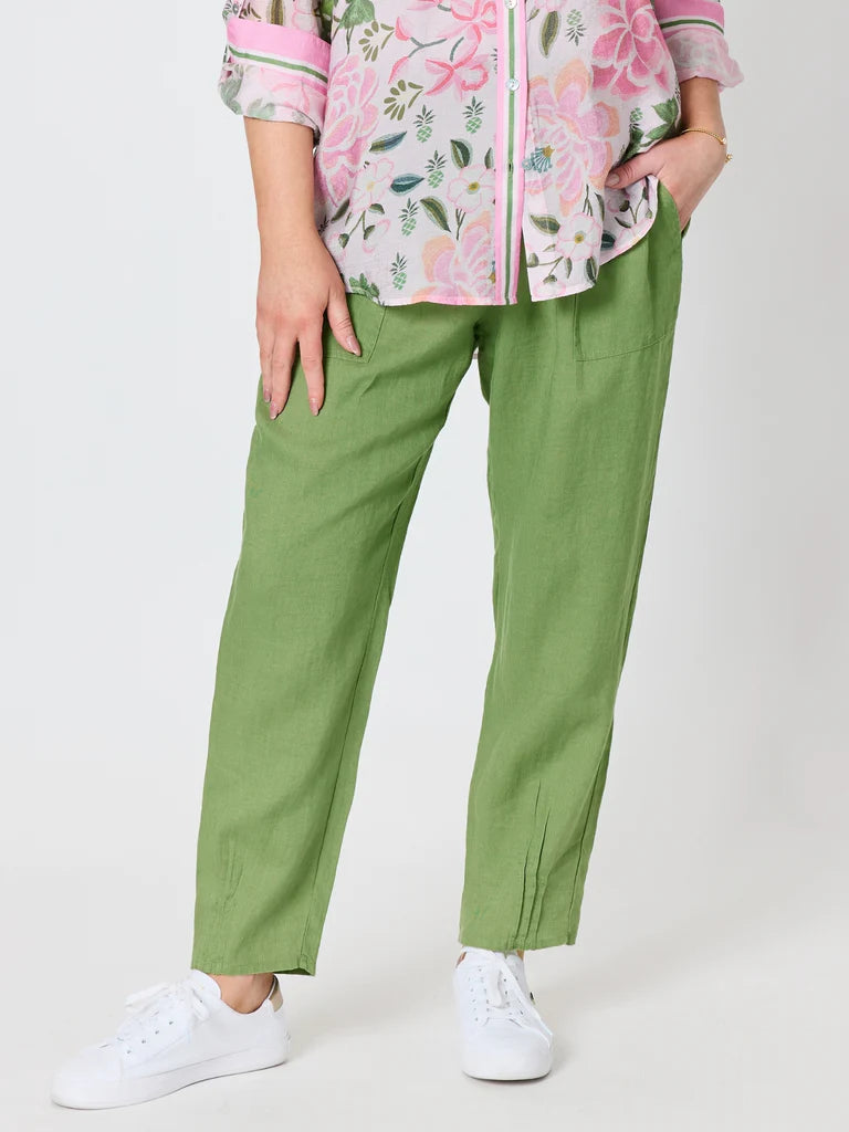 Image of A woman wearing Gordon Smith olive green linen trousers, a pink floral button-up shirt, and white sneakers, standing against a light gray studio background.