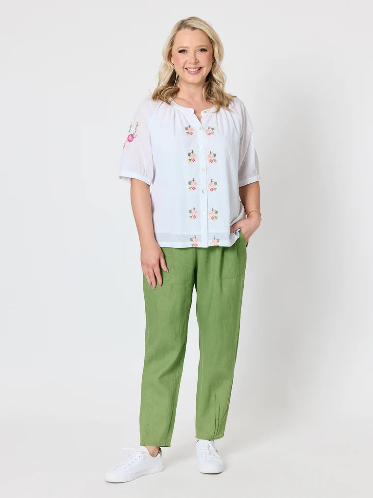 Image of A woman modeling a white short-sleeve blouse with pink floral embroidery and Gordon Smith green trousers, standing against a light gray studio background.