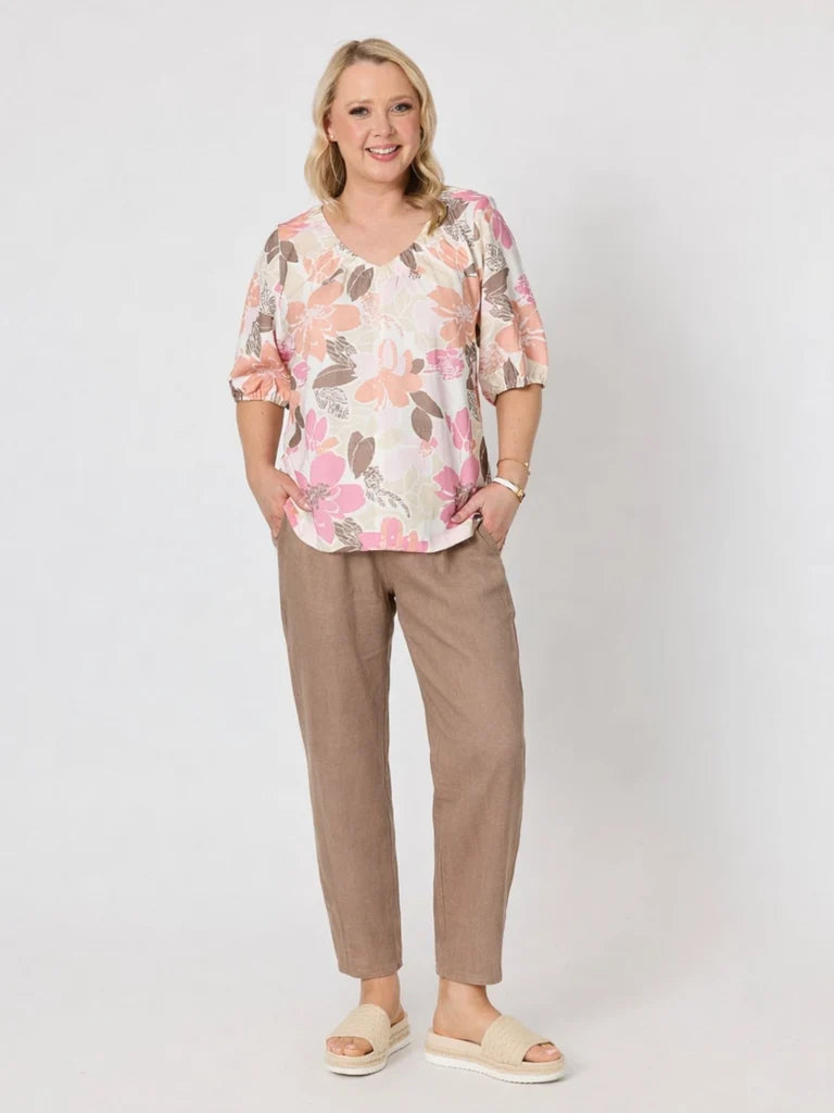 A woman modeling a short-sleeve floral blouse with a V-neck and Gordon Smith brown cropped trousers, standing against a light gray studio background.