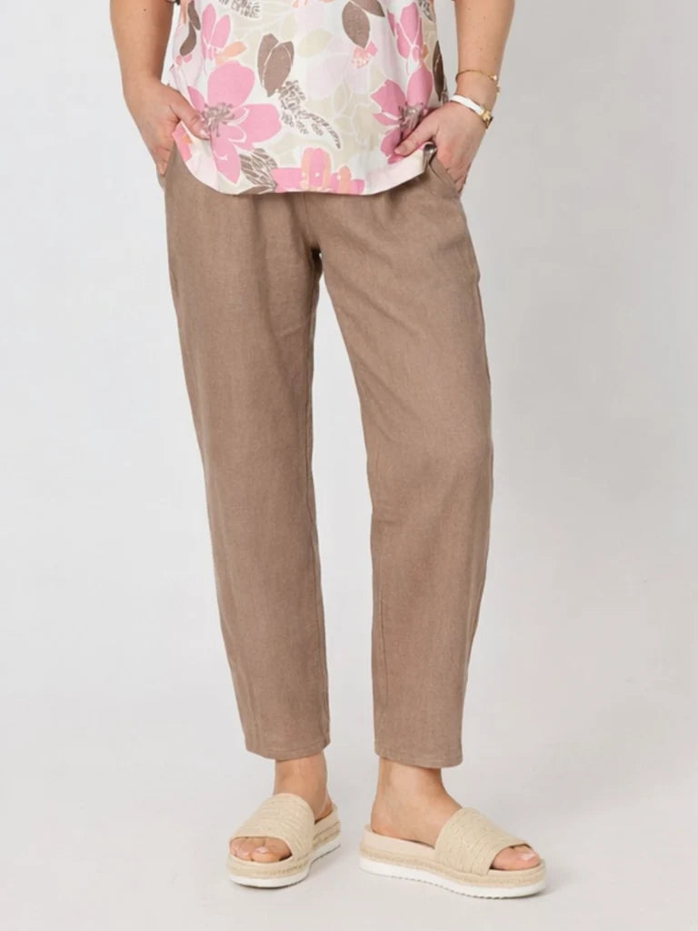 A woman wearing Gordon Smith slim brown trousers, a floral top, and beige slide sandals, standing against a light gray studio background.