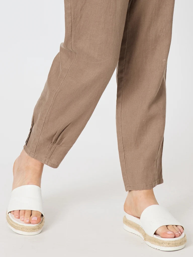 Close-up of a woman wearing Gordon Smith brown linen trousers and white espadrille slide sandals against a light gray studio background.