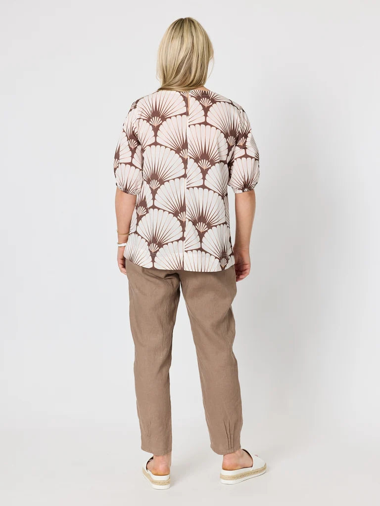A woman viewed from behind wearing a short-sleeve button-up shirt with a brown and cream palm leaf pattern, paired with Gordon Smith brown trousers and white slide sandals, set against a plain light gray studio background.