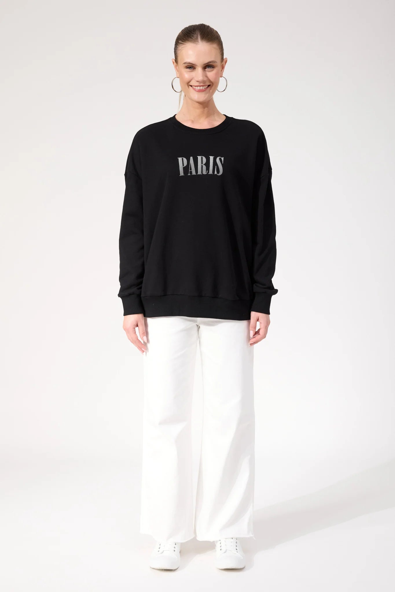 Woman wearing Haven black crewneck paired with white pants on a plain background 