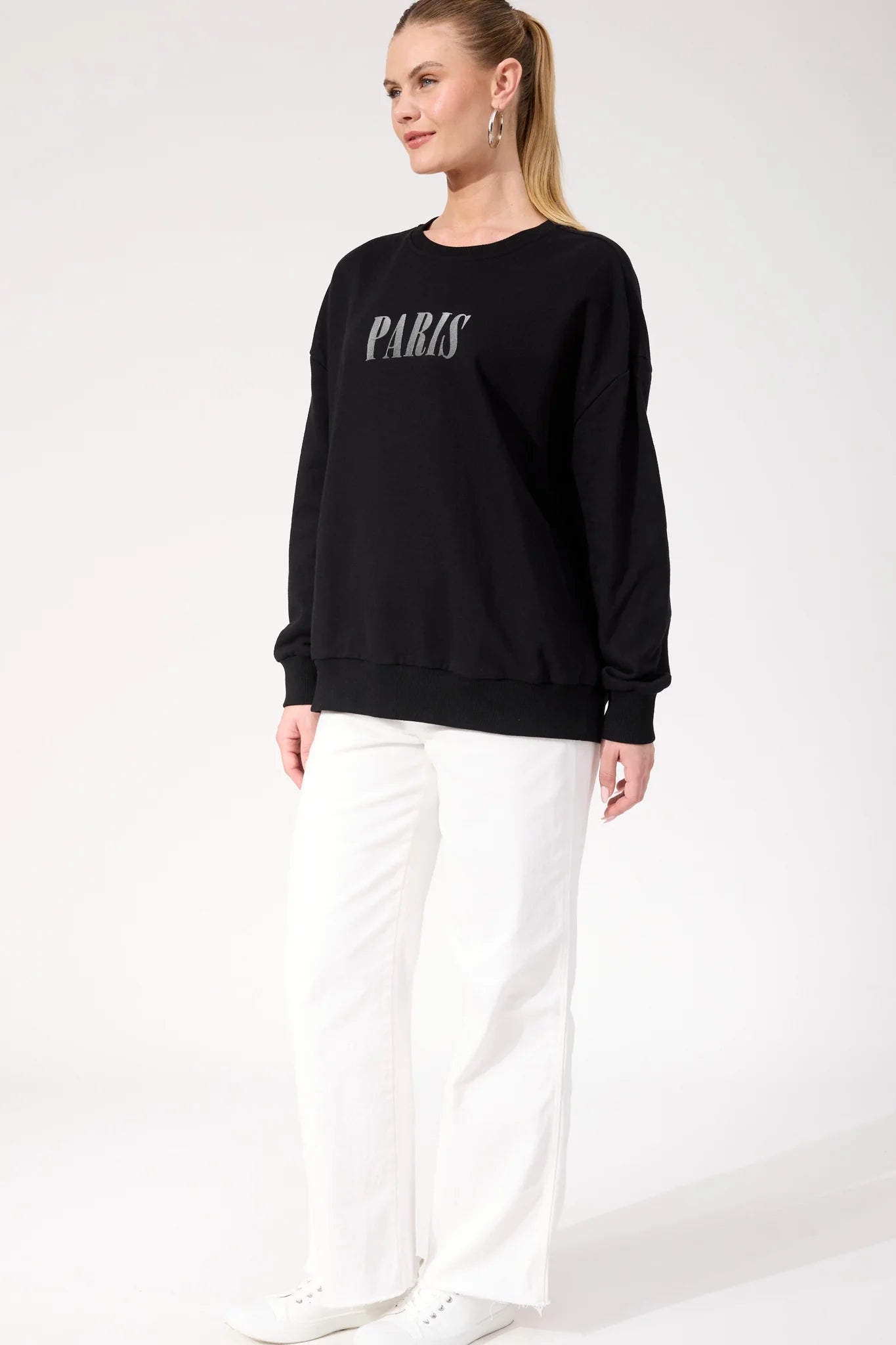 Woman wearing Haven black crewneck paired with white pants on a plain background 