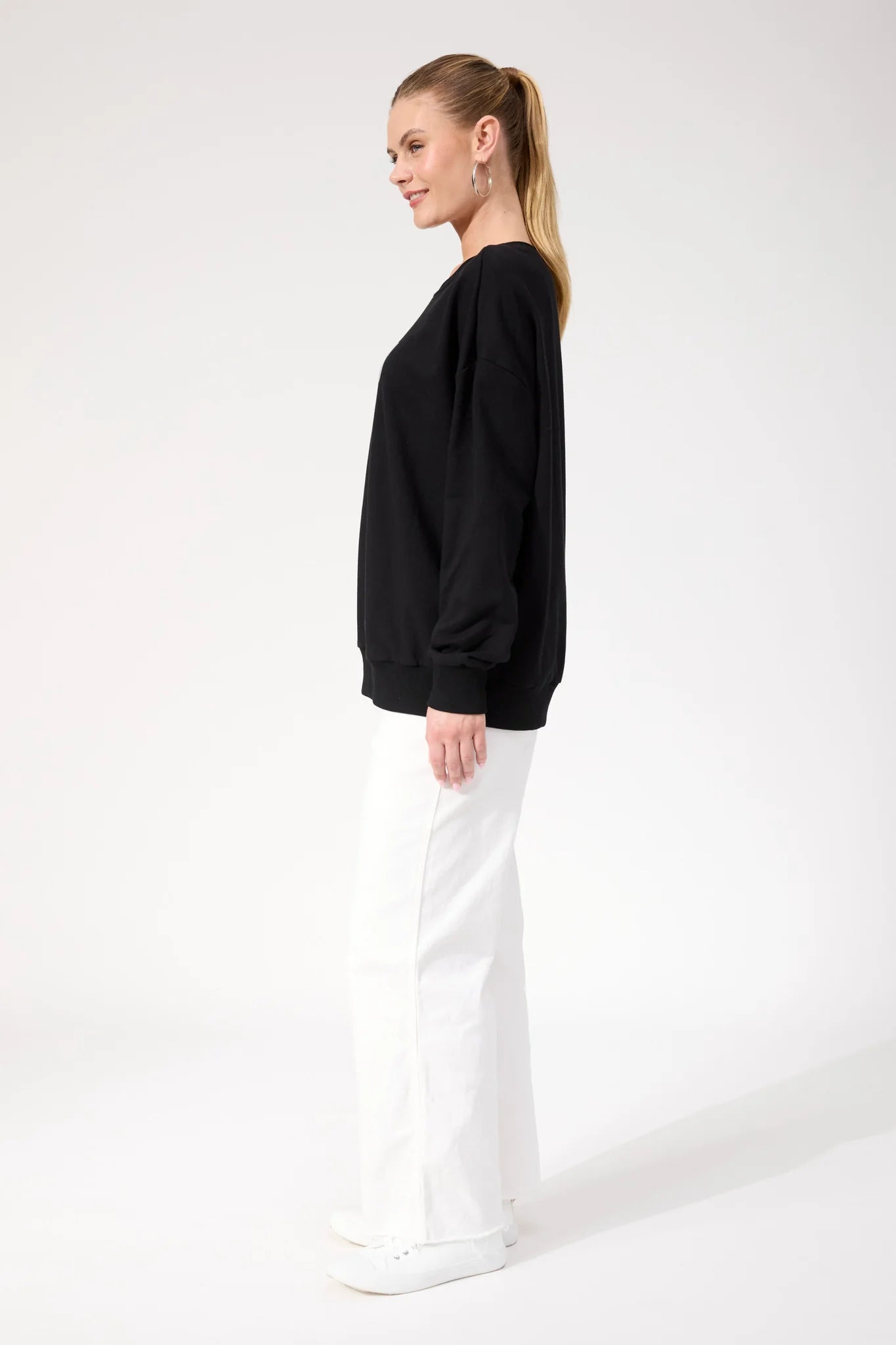 Woman wearing Haven black crewneck paired with white pants on a plain background 