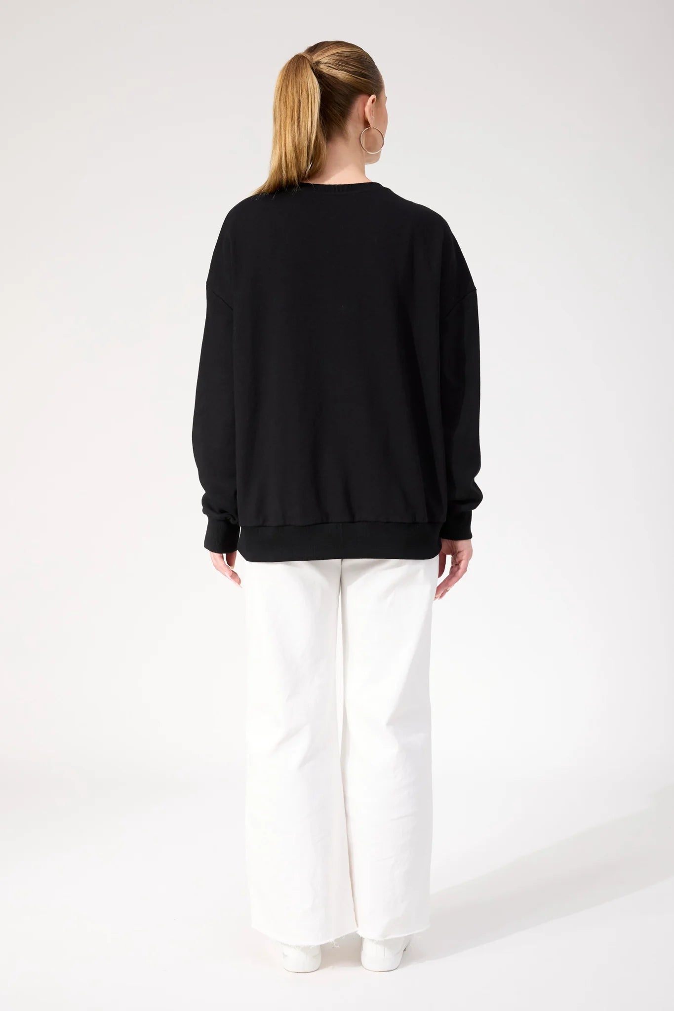 Woman wearing Haven black crewneck paired with white pants on a plain background 