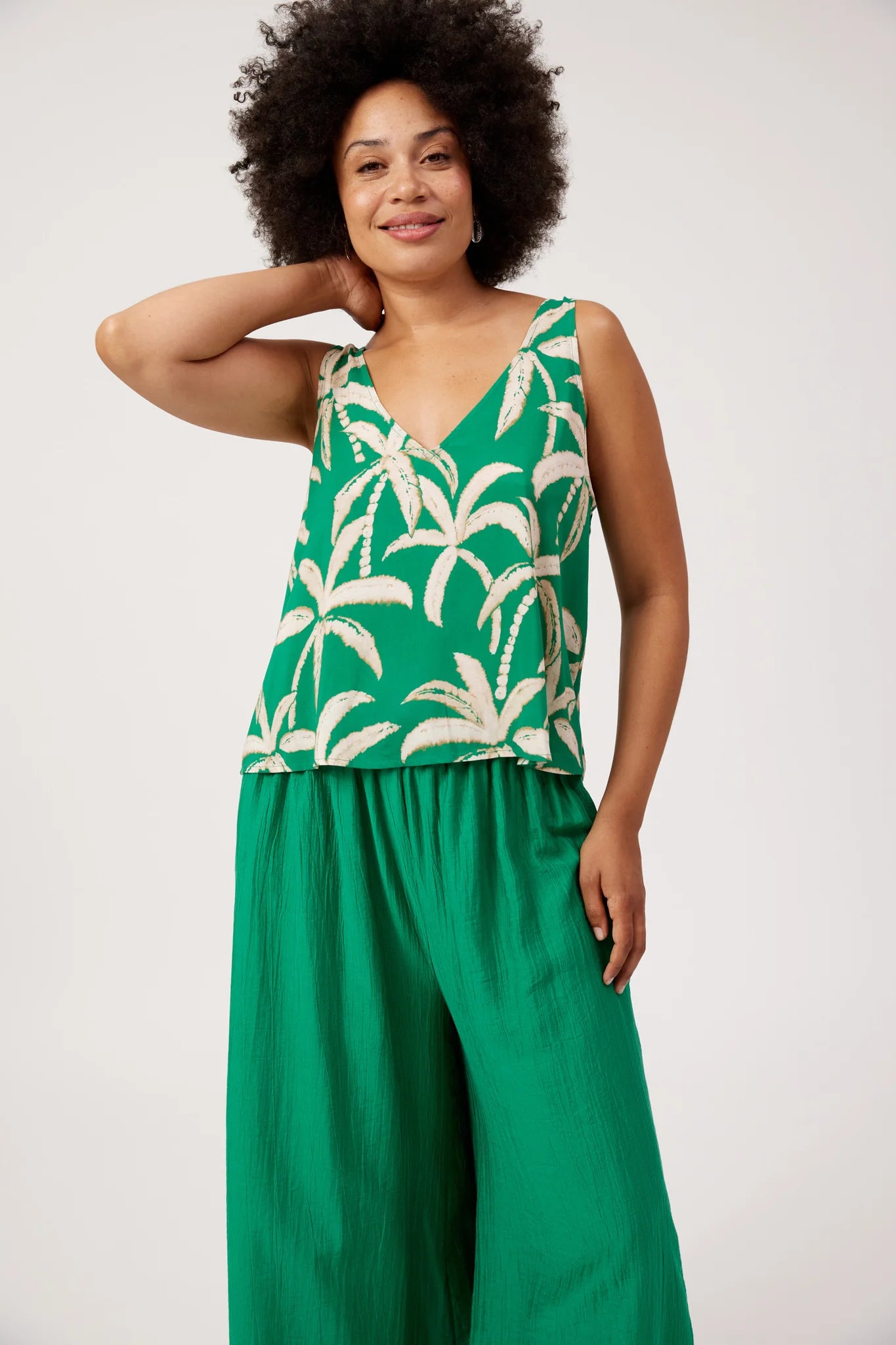 Image of A woman modeling a Haven sleeveless green tank with a cream palm tree pattern on the top and solid green wide-leg pants, photographed against a plain light gray studio background.