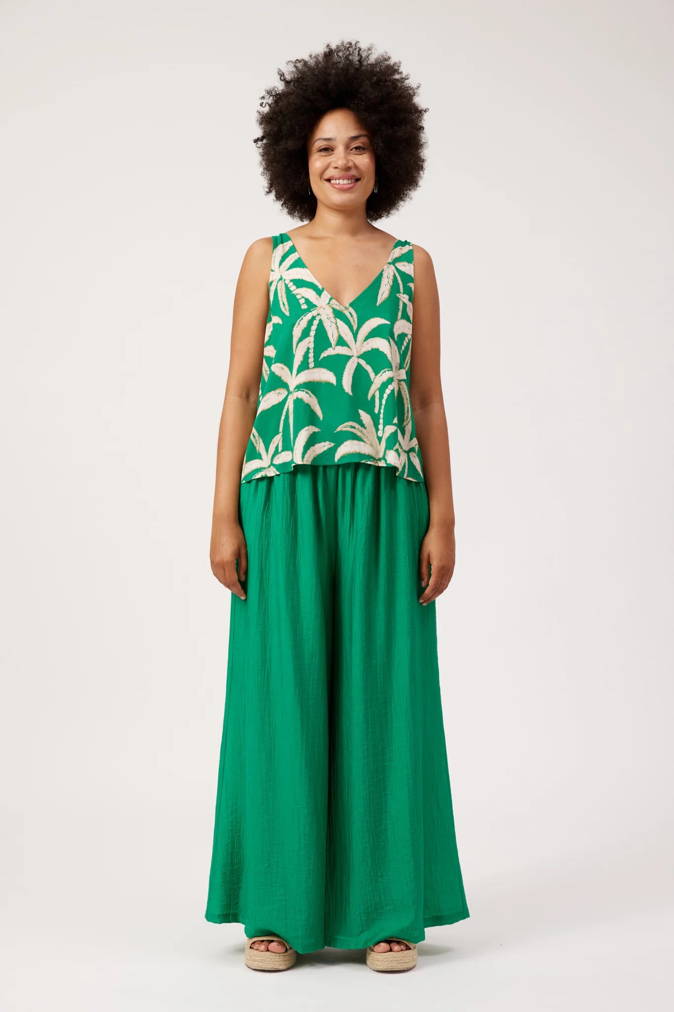 Image of A woman modeling a Haven sleeveless green maxi dress with a white palm tree pattern on the bodice and solid green wide-leg skirt, standing against a plain white studio background.