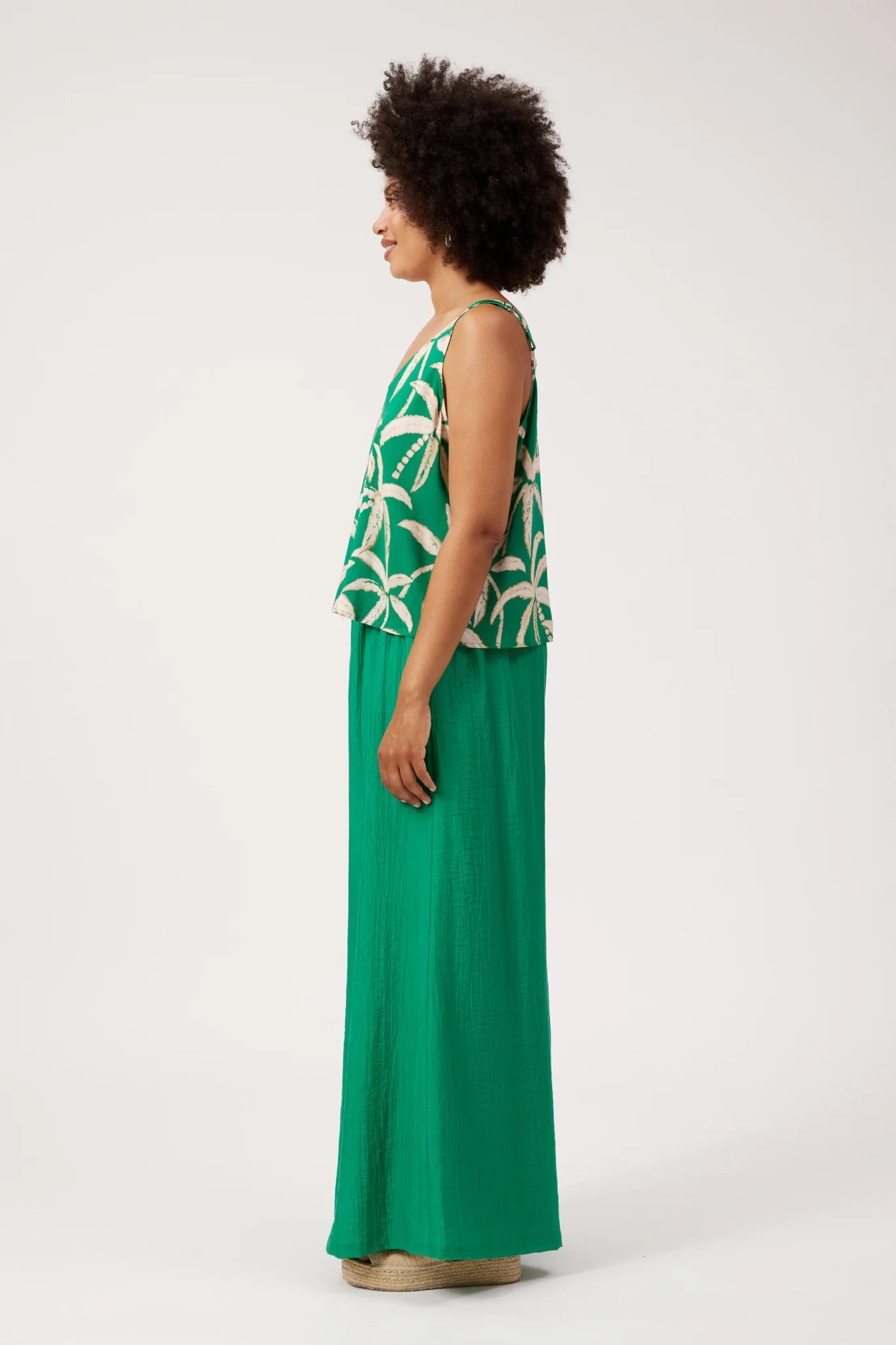 Image of A woman shown in side profile wearing a Haven sleeveless green top with a white palm leaf pattern and a matching long green skirt, standing against a plain light gray studio background.