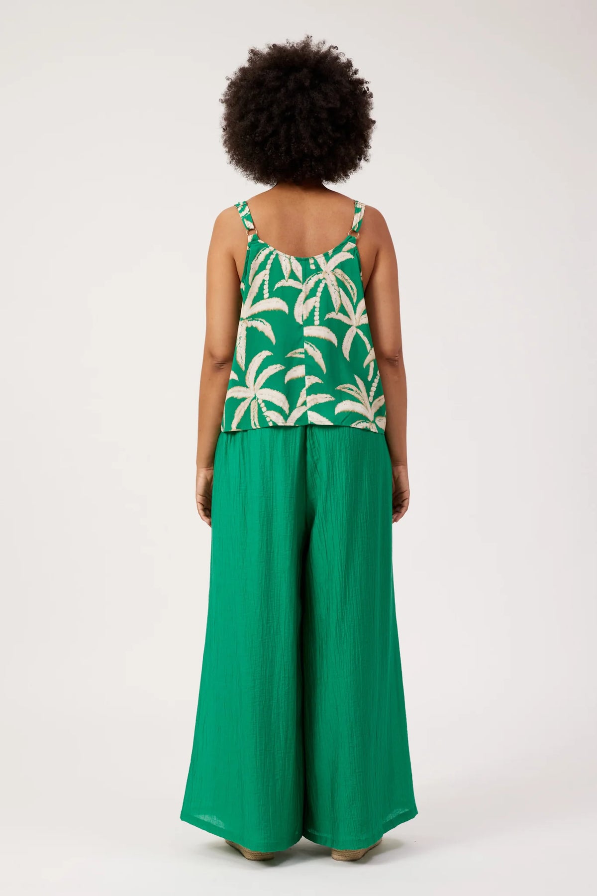A model viewed from the back wearing a Haven green sleeveless top with a cream palm tree pattern and matching green wide-leg pants, set against a plain light gray studio background.