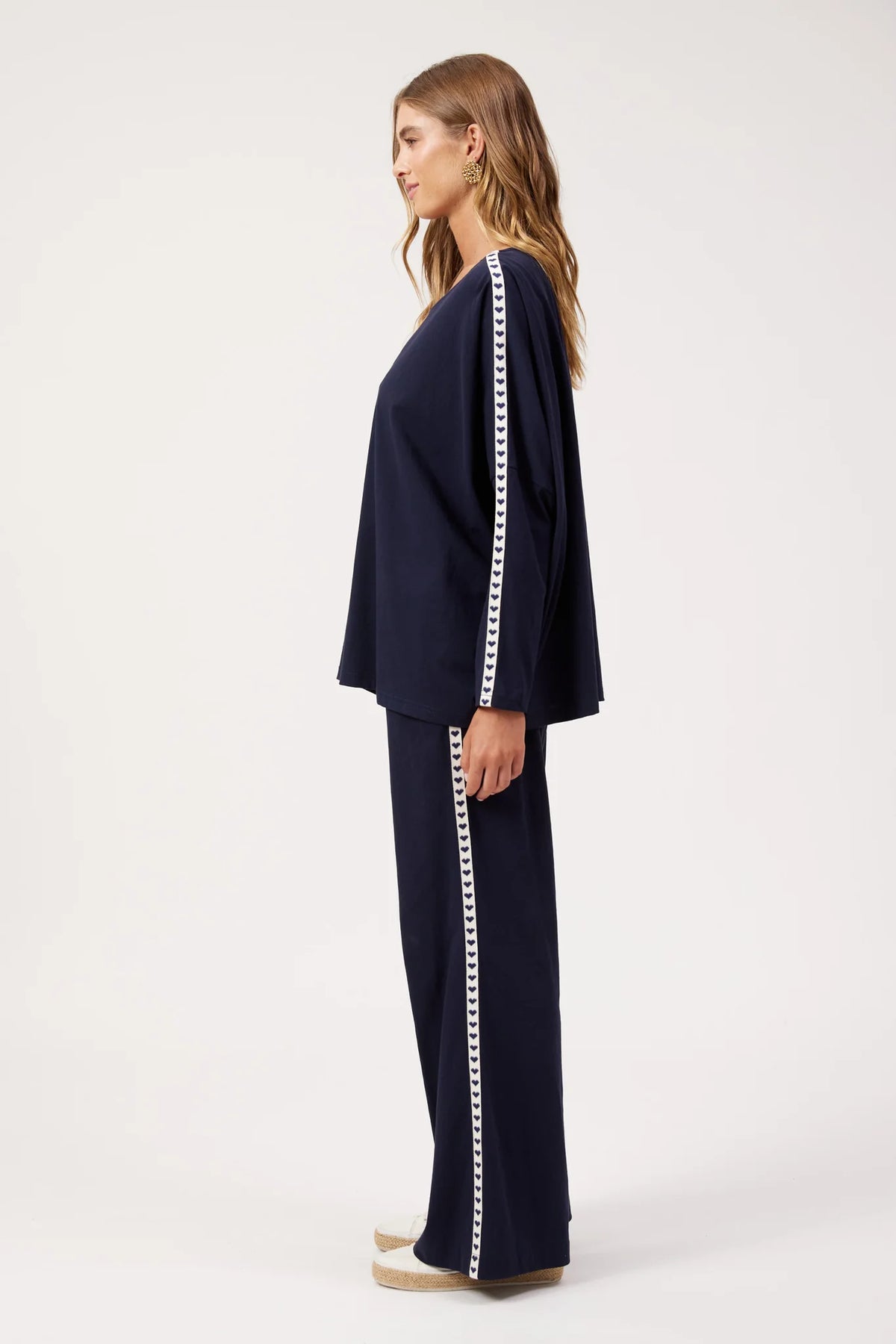 A woman stands in profile wearing a Haven navy blue lounge set with a matching top and wide-leg pants, both featuring a white decorative stripe down the side, photographed against a plain light gray studio background.