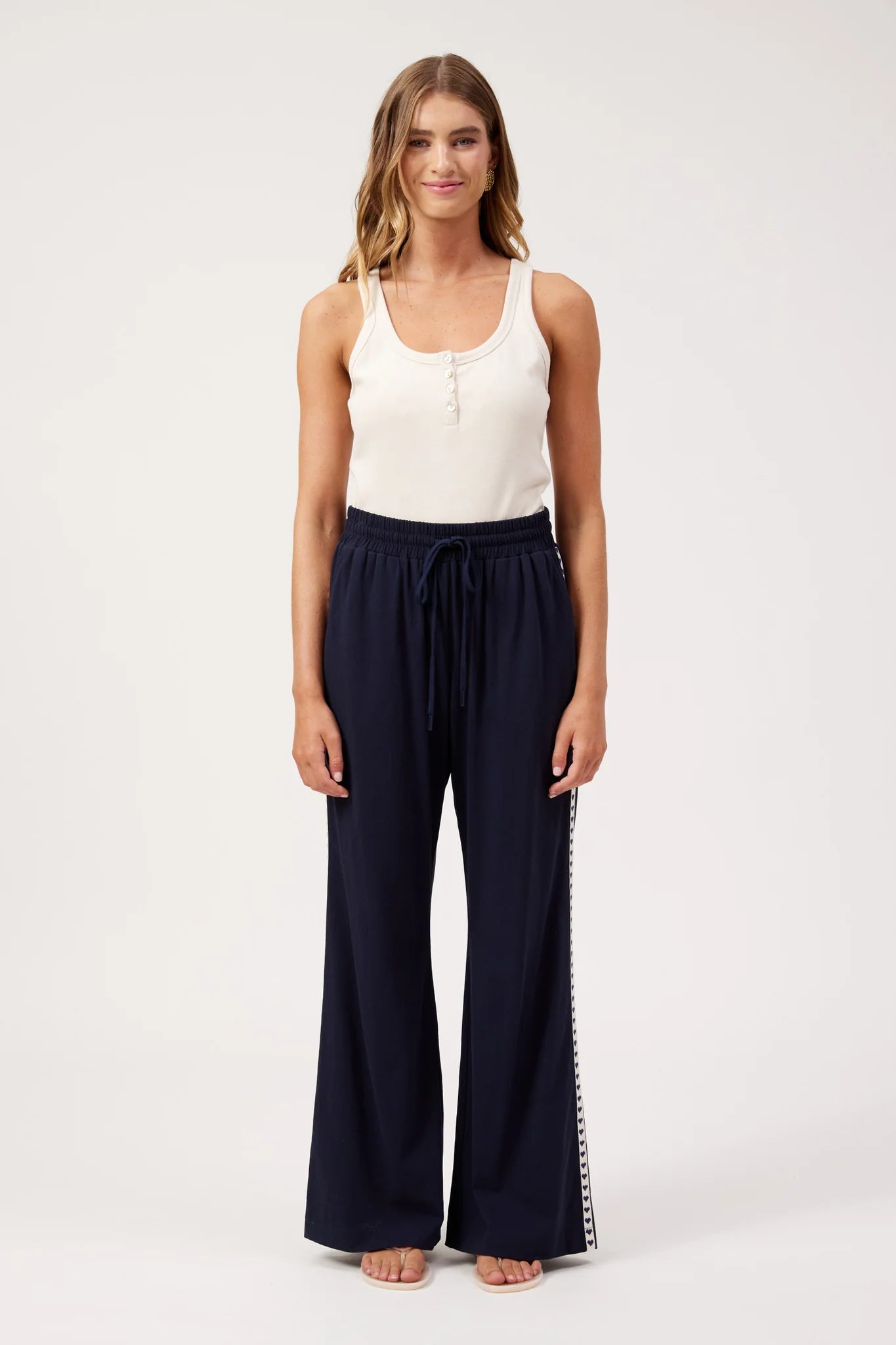 Image of A woman modeling a pair of Haven dark navy wide-leg pants with a side stripe detail, paired with a white sleeveless top, against a plain white studio background.