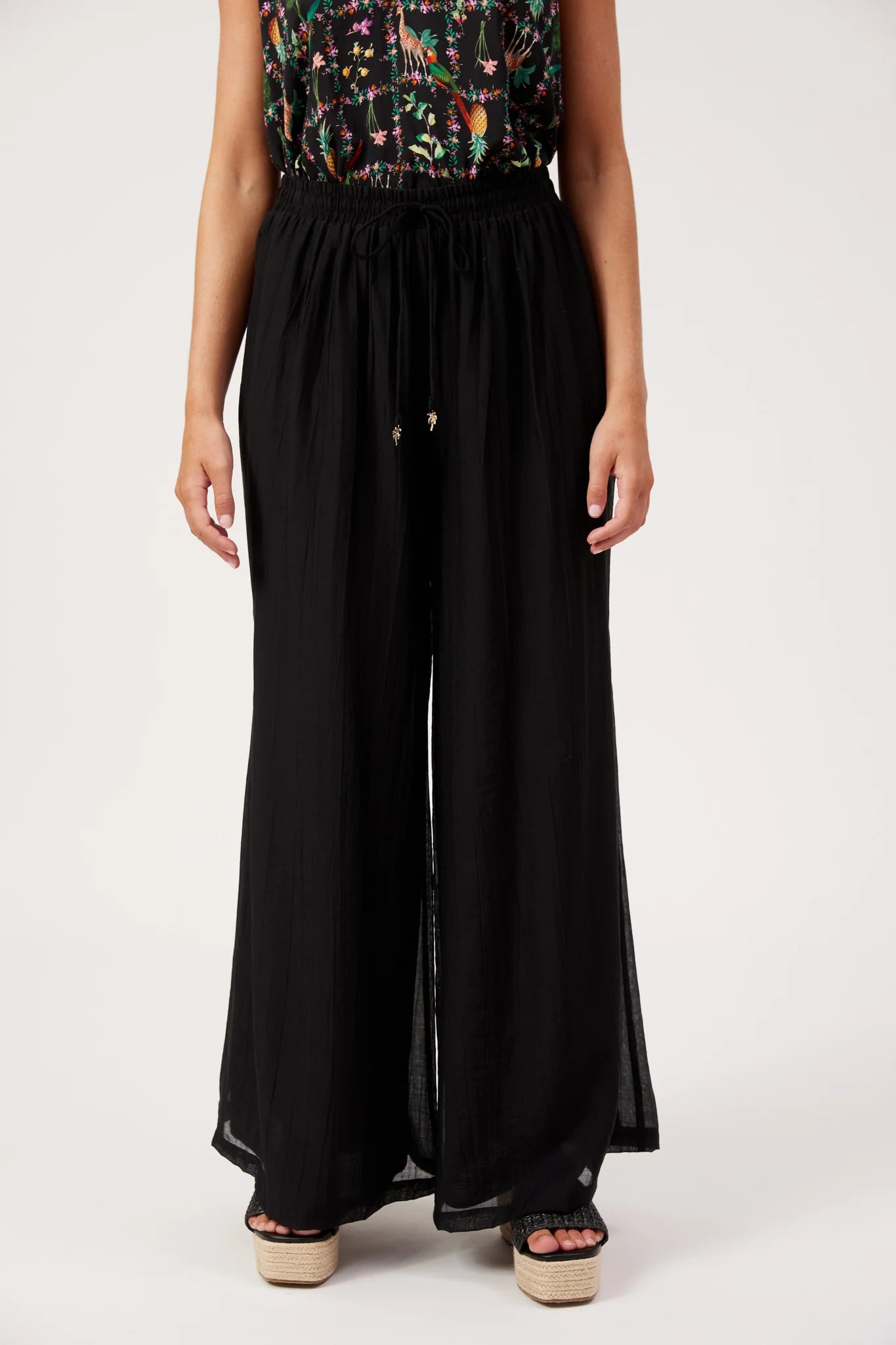 Image of A model wearing Haven black wide-leg pleated trousers with a drawstring waist, paired with a colorful floral sleeveless top and black platform sandals, against a plain white studio background.