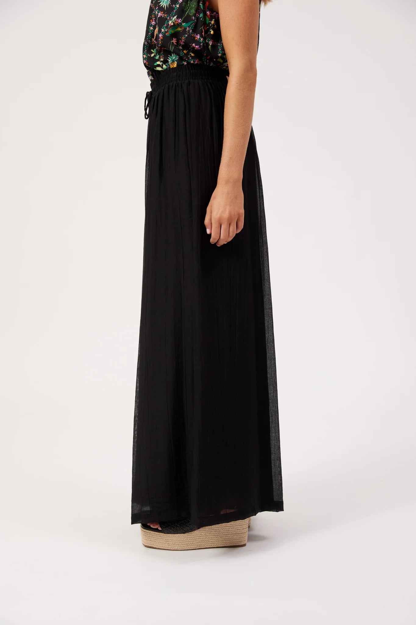 Image of A model is shown from the side wearing a Haven long black pleated maxi skirt with a sheer side panel and a black waist tie, paired with a colorful floral top and platform sandals against a white studio background.