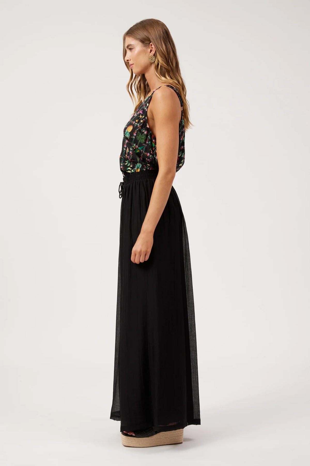 A woman stands in profile wearing a sleeveless top with a Haven long black pants, photographed against a plain light gray studio background.