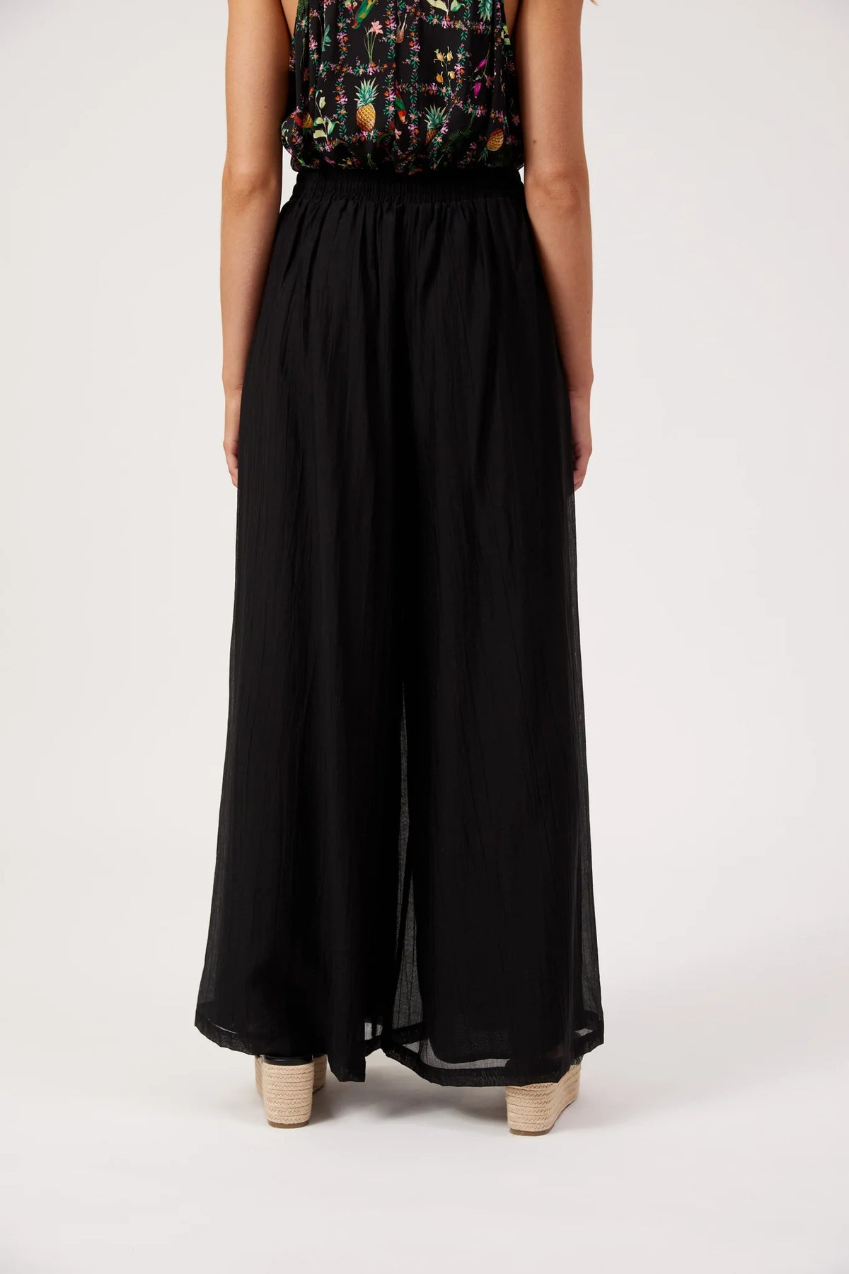 A model from the back wearing Haven black wide-leg pleated trousers and a colorful patterned sleeveless top, standing against a plain light gray studio background.