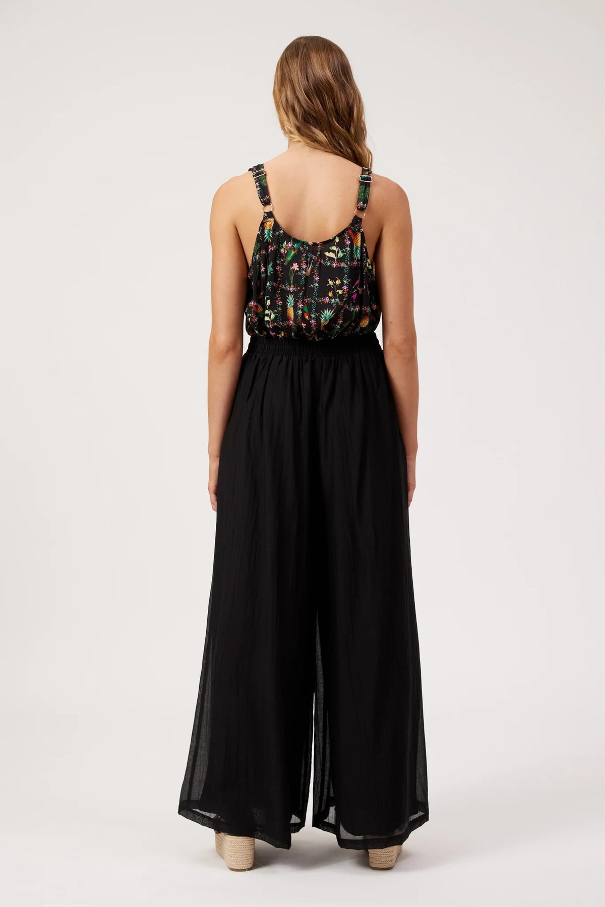 A woman viewed from the back wearing a Haven sleeveless black wide-leg jumpsuit with a multicolor patterned bodice, standing against a plain light gray studio background.
