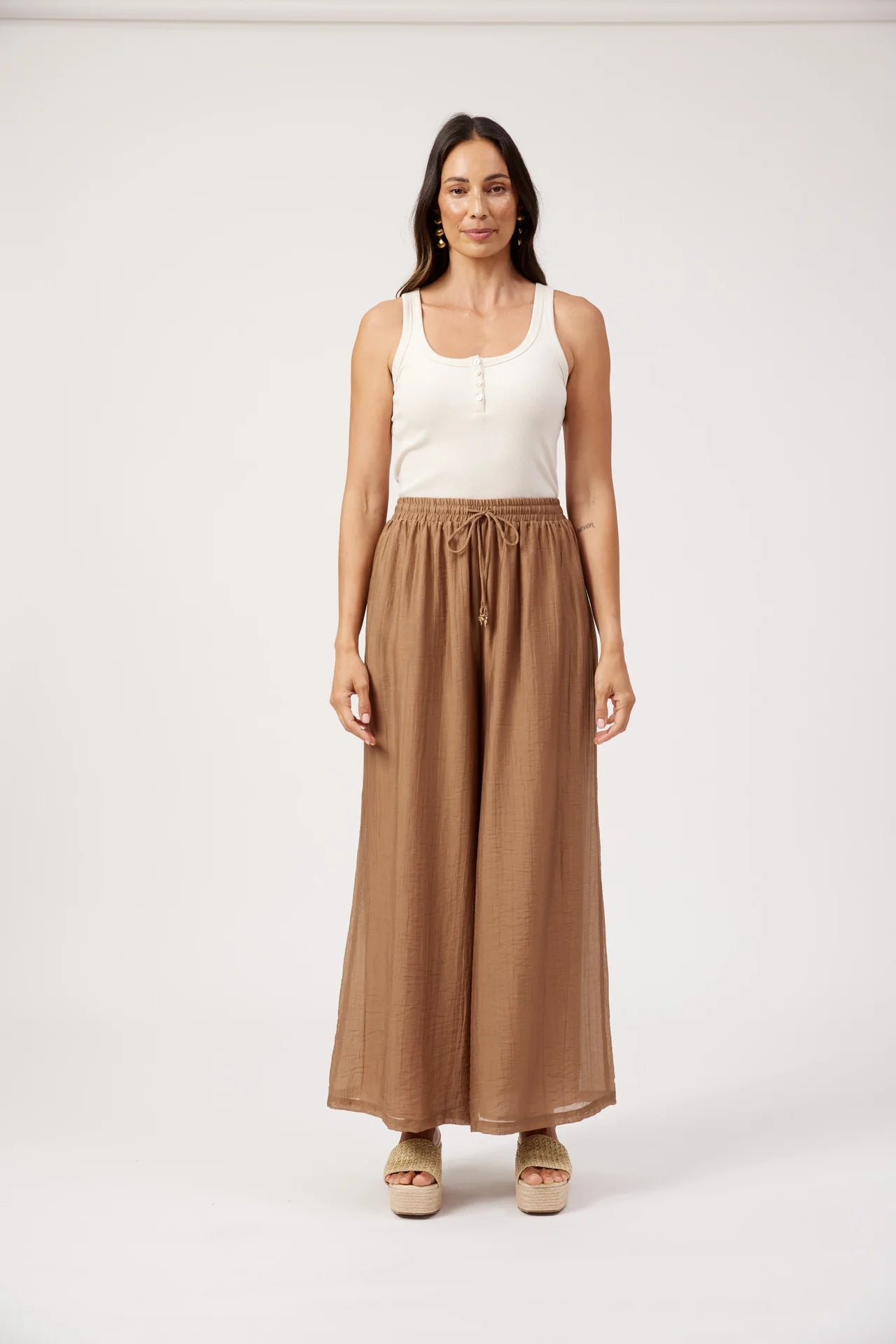 Image of A woman stands against a plain light gray studio background wearing a white sleeveless top and Haven high-waisted, wide-leg brown pants with a drawstring waist.