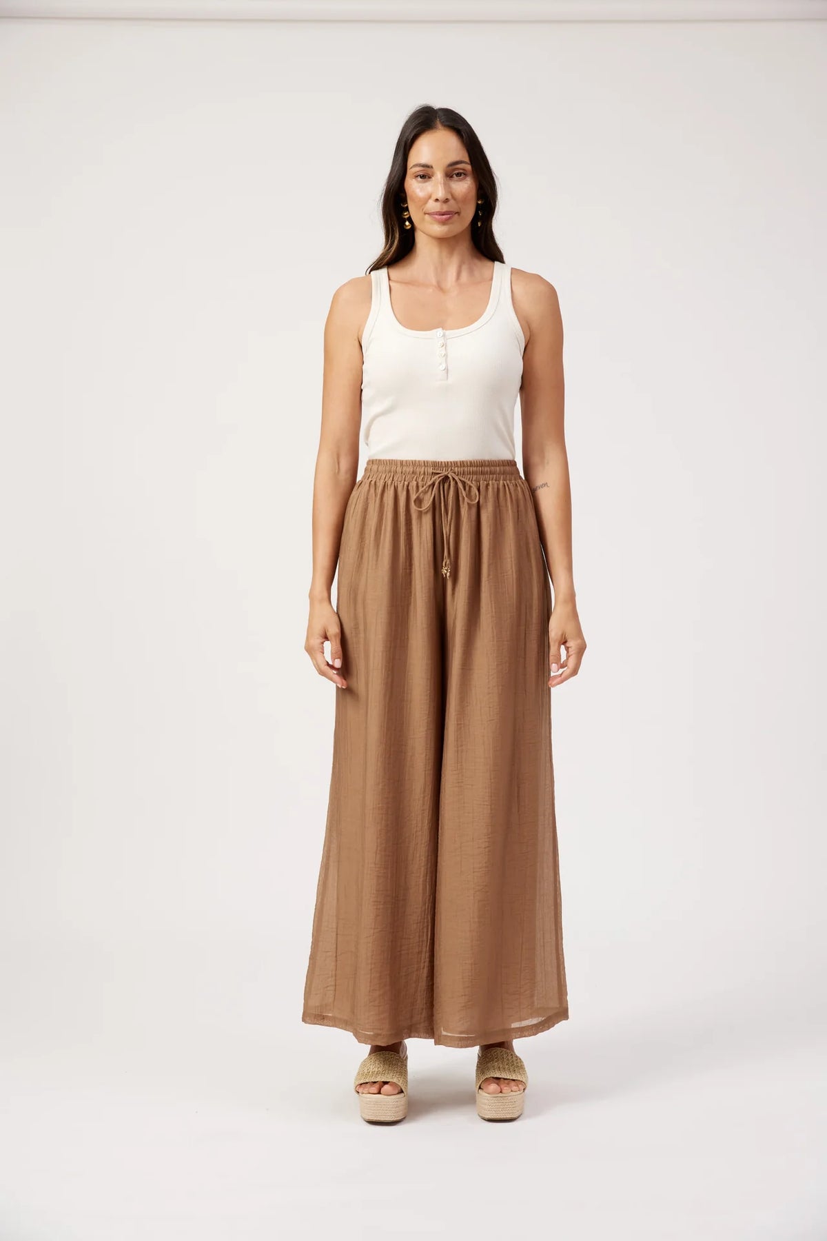 A woman stands against a plain light gray studio background wearing a white sleeveless top and Haven high-waisted, wide-leg brown pants with a drawstring waist.