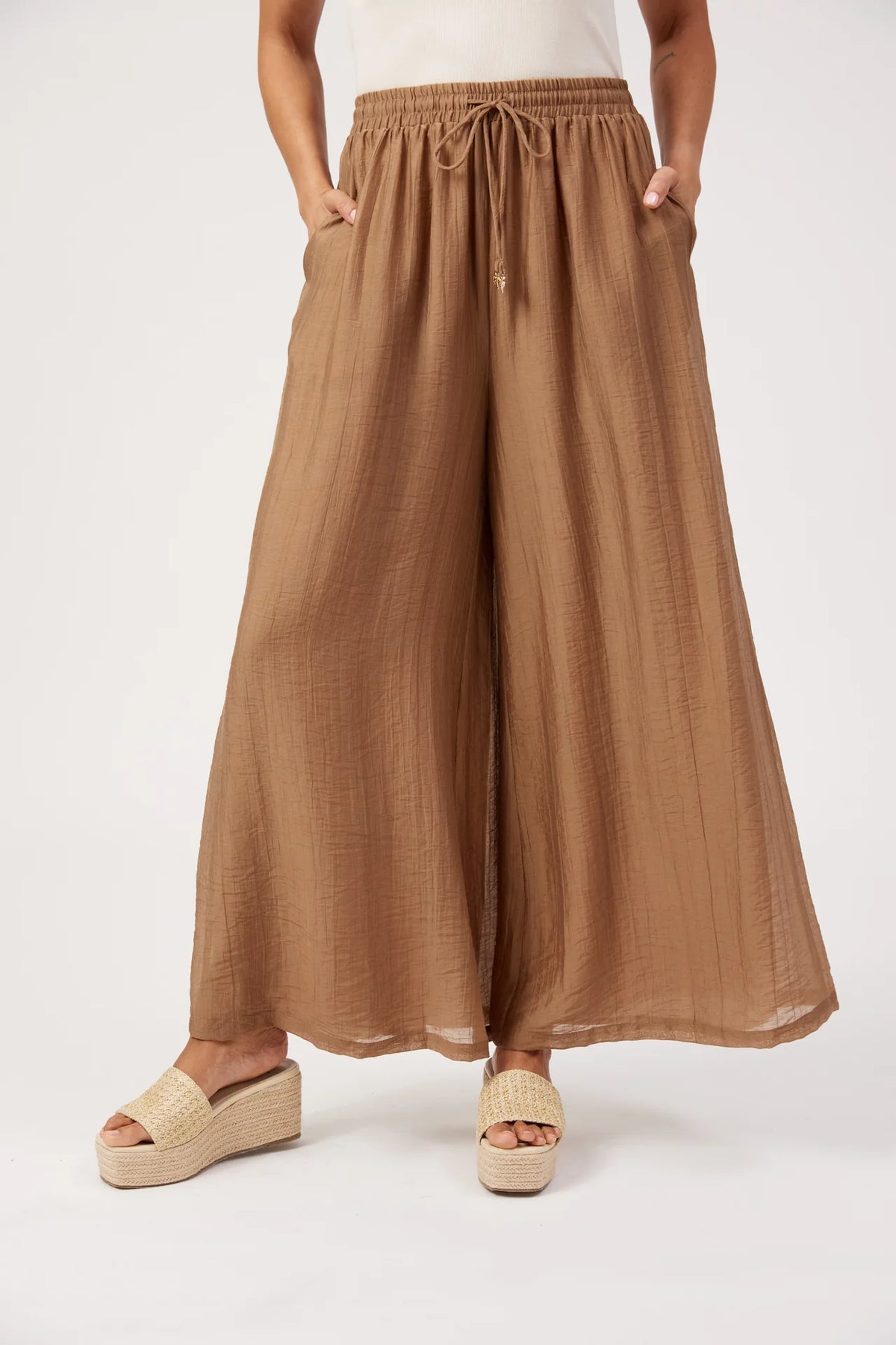 A woman Haven wearing loose, wide-leg pleated pants in a warm tan/camel color, paired with beige platform sandals, shown against a plain light gray studio background.