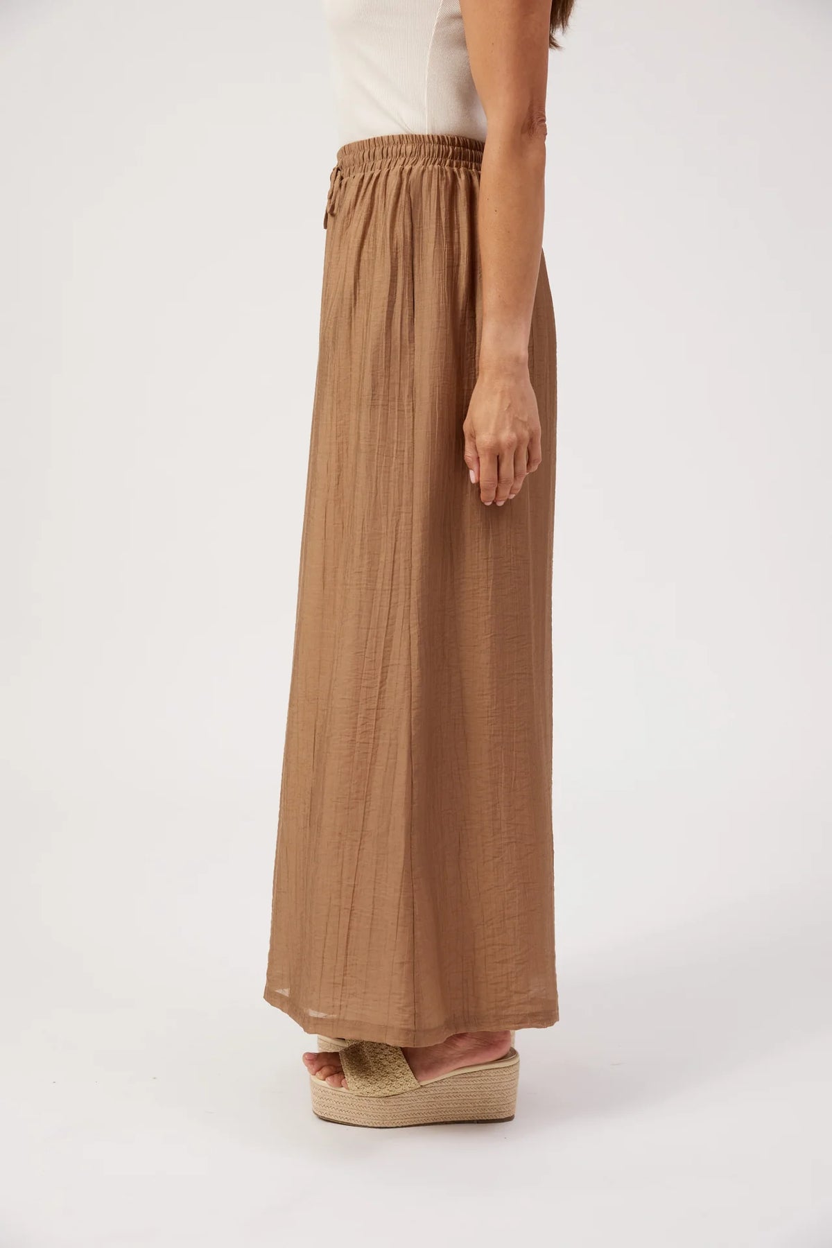 A woman shown from the waist down wearing a Haven long, wide-leg brown skirt with an elastic waistband, paired with a white top and espadrille wedge sandals, against a plain white studio background.