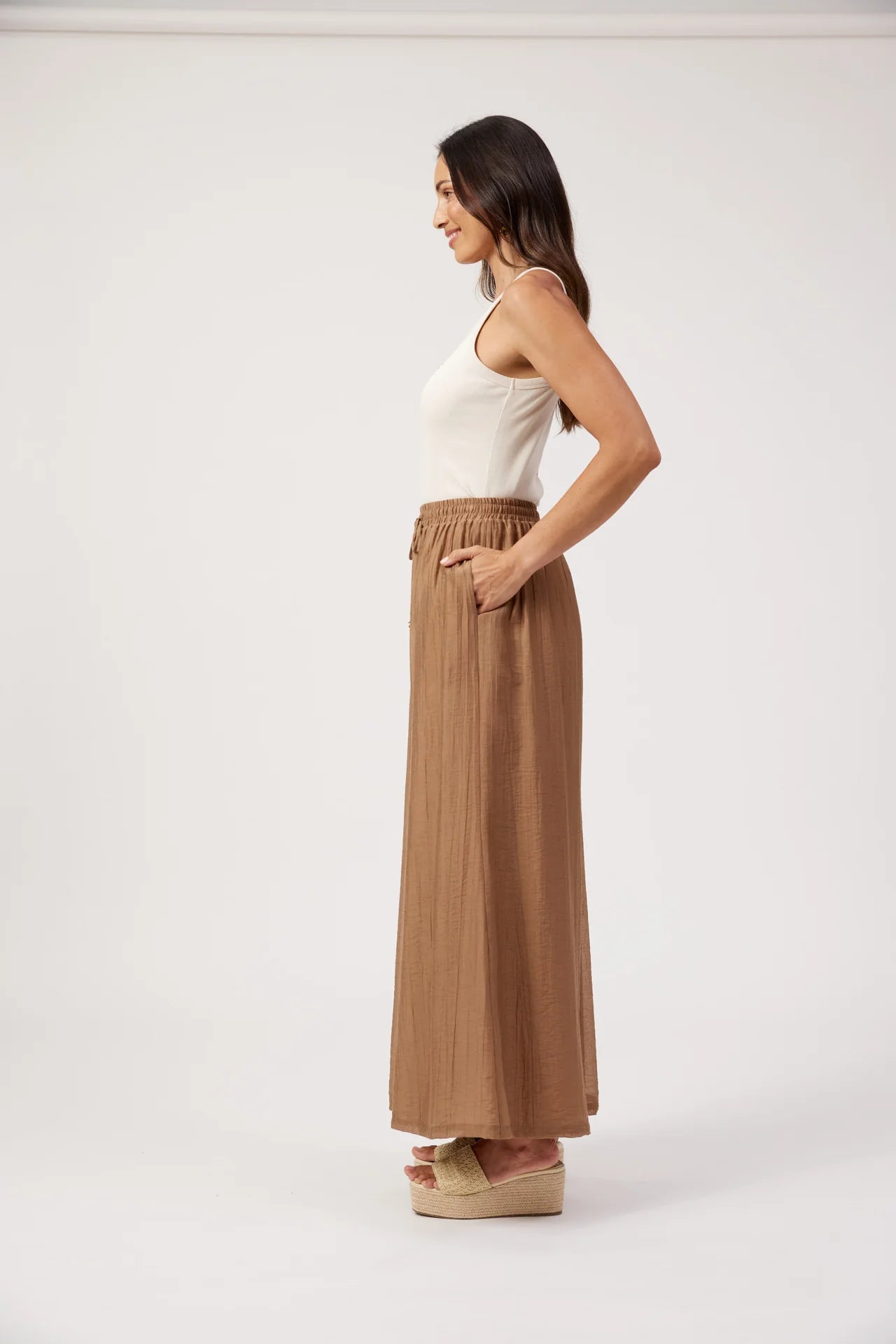 Image of A woman stands in profile wearing a white ribbed tank top and Haven long, pleated, high-waisted pants in a warm taupe-brown color, paired with beige platform wedge sandals, against a plain white studio background.