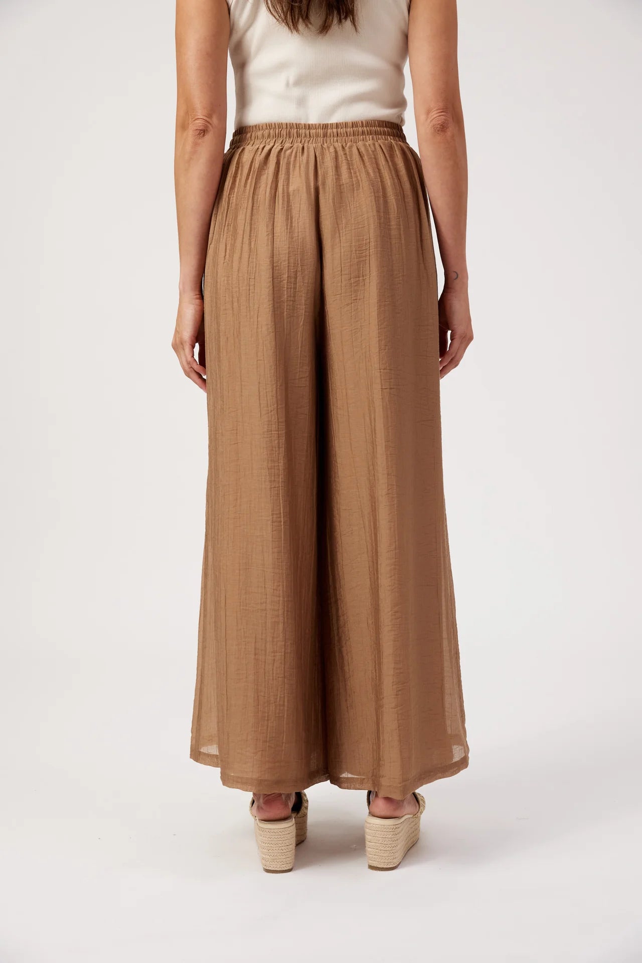 Image of A model shown from the back wearing Haven high-waisted, wide-leg cropped pants in a warm tan or camel color, paired with a cream sleeveless top and beige platform sandals, against a plain white studio background.