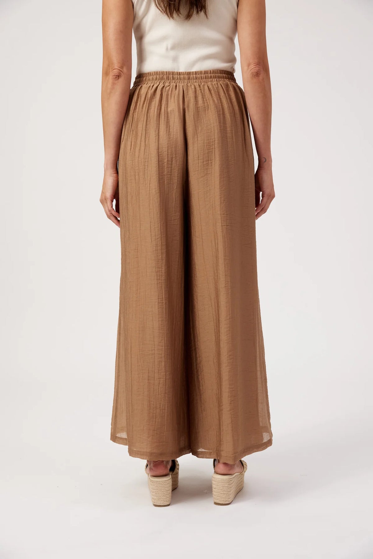 A model shown from the back wearing Haven high-waisted, wide-leg cropped pants in a warm tan or camel color, paired with a cream sleeveless top and beige platform sandals, against a plain white studio background.
