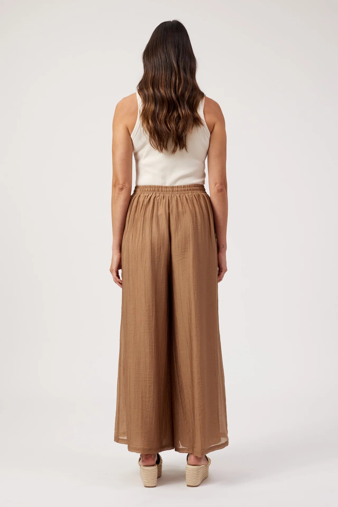 Image of A woman shown from the back wearing a white sleeveless top and Haven high-waisted, wide-leg brown pants, standing against a plain light gray studio background.