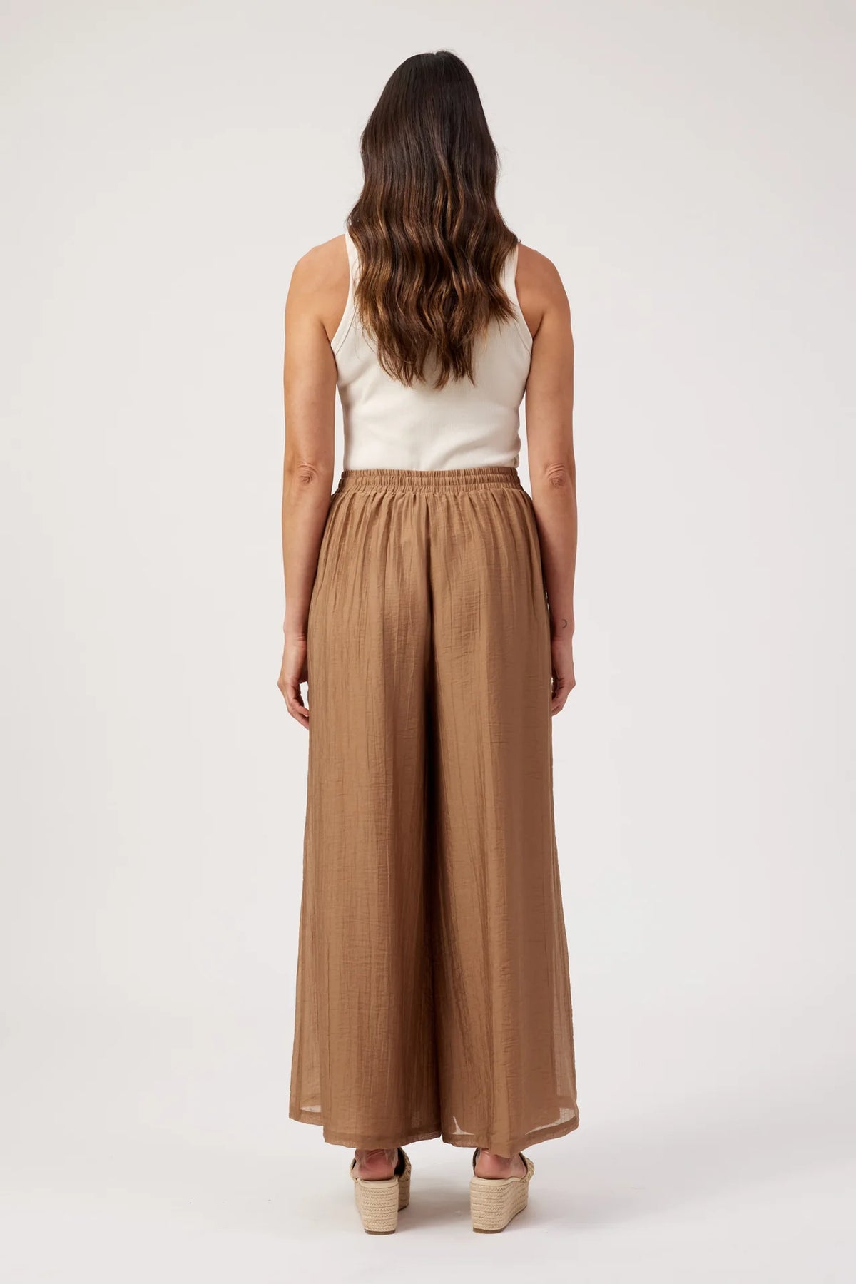 A woman shown from the back wearing a white sleeveless top and Haven high-waisted, wide-leg brown pants, standing against a plain light gray studio background.