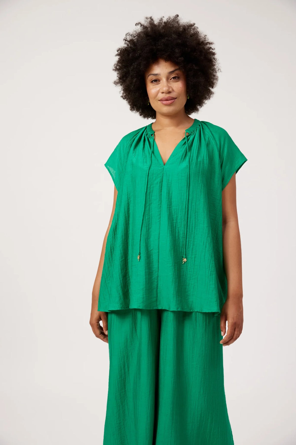 A woman with an afro hairstyle wearing a Haven bright green two-piece matching set consisting of a short-sleeve V-neck top with drawstrings and wide-leg pants, photographed against a plain light gray studio background.