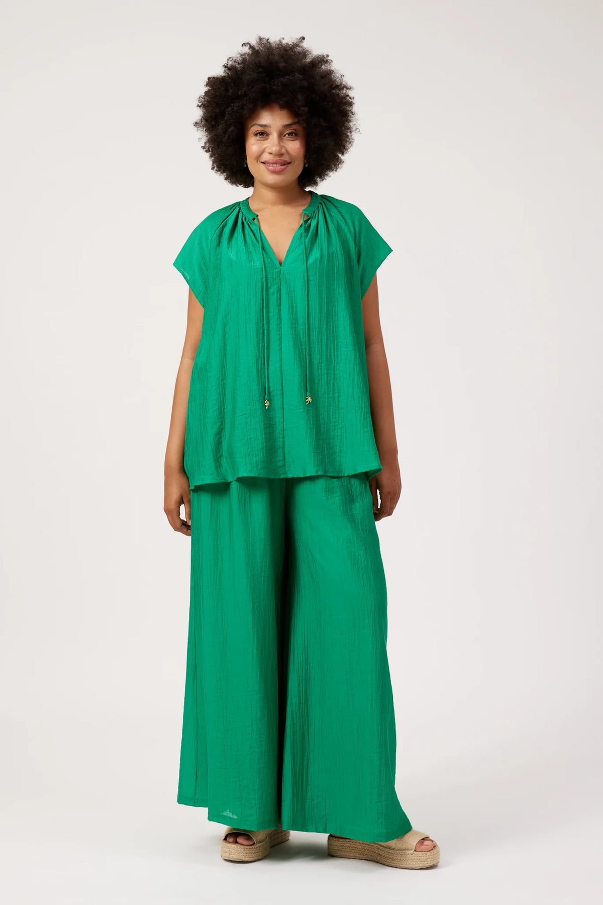 A woman modeling a Haven matching green two-piece outfit consisting of a short-sleeve V-neck top with drawstrings and wide-leg pleated pants, photographed against a plain light gray studio background.