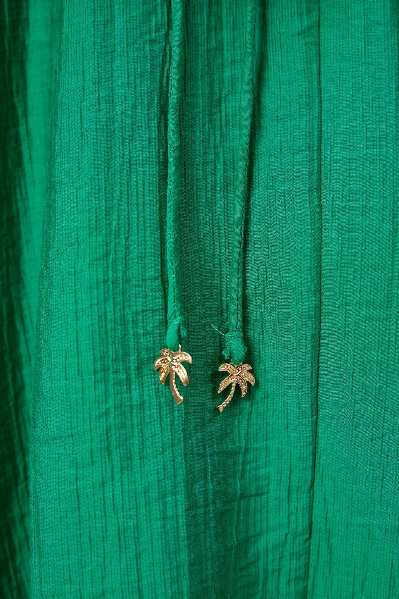 Image of Close-up of a Haven textured green fabric garment with two drawstrings ending in small gold palm tree charms.